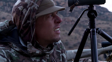 Hunter in camouflage hoodie looking up beside tripod-mounted spotting scope