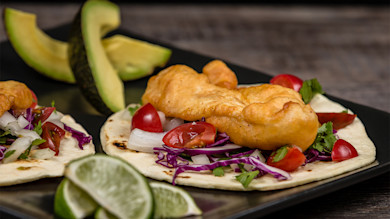 Two fish tacos with battered fish, purple cabbage, cherry tomatoes, onions, lime wedges, avocado