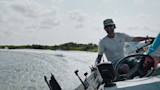 Man steering boat through choppy marsh water, smiling, wearing cap and sunglasses