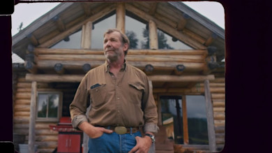 Bearded man standing with hands in pockets in front of a log cabin