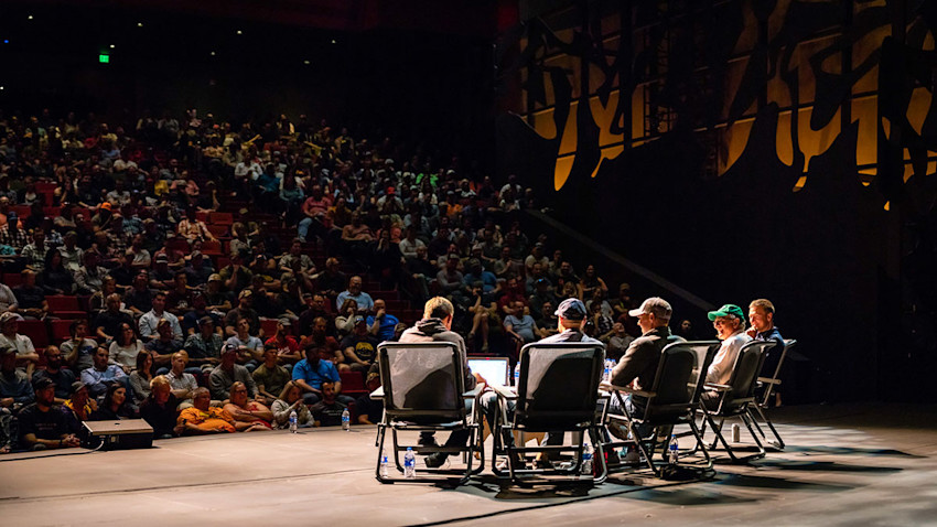 Panel of five speakers seated on stage facing a large theater audience