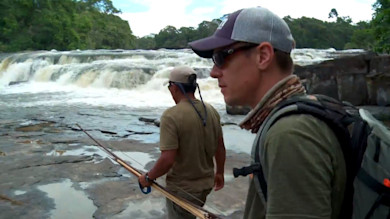 Two men on rocky riverbank by waterfall carrying long fishing poles