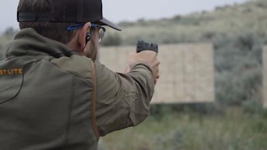 Man aiming a pistol at an outdoor target, wearing a First Lite jacket and cap