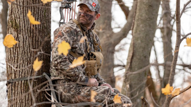 Hunter in camouflage and face paint seated on a tree stand with bow in autumn woods