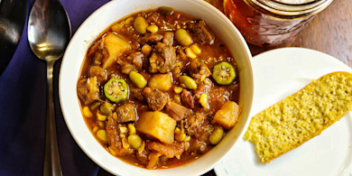Hearty stew in bowl with okra, corn, potatoes and chunks of meat; spoon and cracker on plate