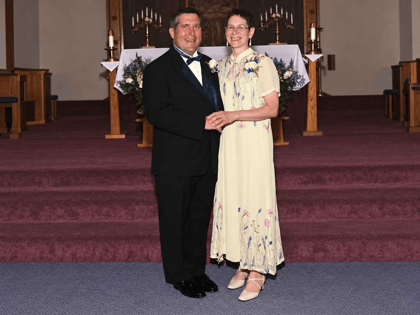 Couple in formal wear holding hands at a church altar with floral arrangements and candles