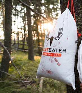 Bloodstained white bag hanging from tree with 'MEATEATER' logo and 'GROCERY BAG' text