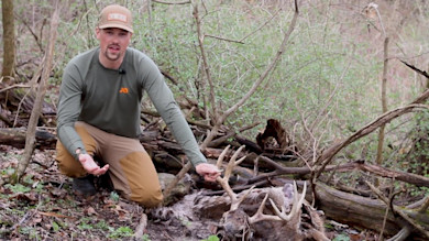 Man kneeling in woods holding a buck's antlers next to a dead deer head; cap reads YETI