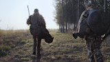 Two hunters walking away toward a tree line; one carries a harvested turkey