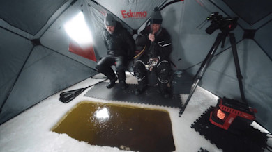 Two anglers inside an Eskimo ice-fishing shelter, one holding a rod over a rectangular hole