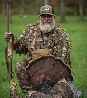 Bearded hunter kneeling with shotgun and turkey tail, wearing cap reading "BEAR GREASE"