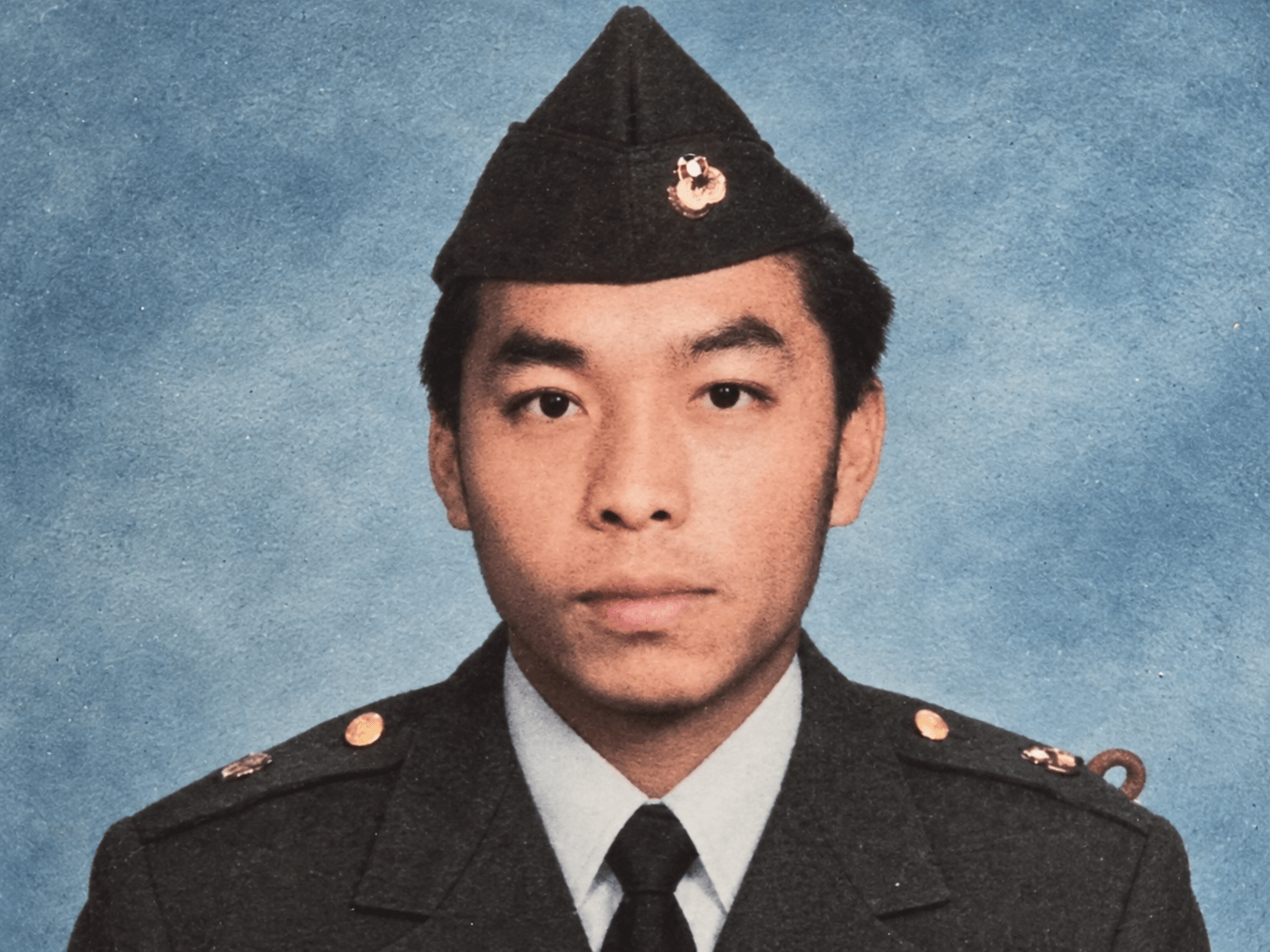 Young man in military dress uniform wearing garrison cap against blue backdrop