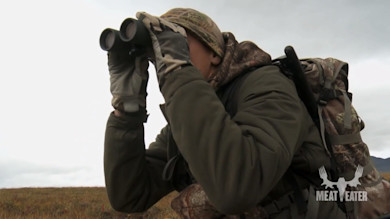 Hunter in camouflage using binoculars with backpack and rifle; MEAT EATER logo