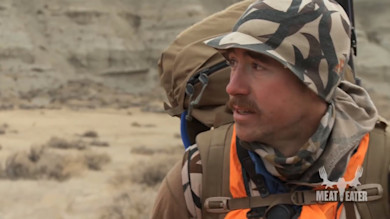 Hunter in camo hat and orange vest carrying backpack; MEAT EATER logo visible