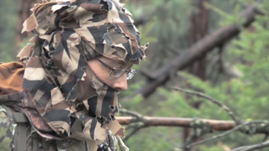Hunter wearing torn camouflage ghillie hood and glasses, side profile in forest