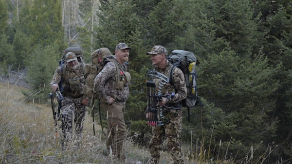 Three hunters in camouflage with large backpacks, one holding a compound bow in pine forest