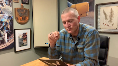 Man seated at desk reading a card with white MeatEater logo; wall sign "MEATEATER"