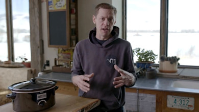 Man explaining venison chili in kitchen, gesturing beside slow cooker labeled WESTON