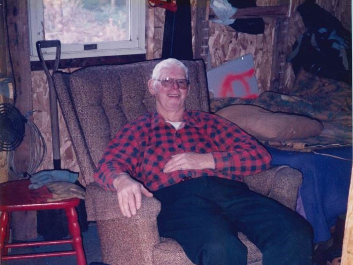 Older man in red plaid shirt and glasses smiling in armchair inside a rustic shed