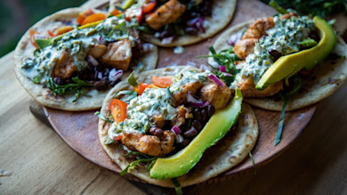 Three grilled fish tacos on a wooden board with avocado, black beans, crema, red onion, tomato