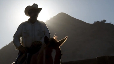 Backlit cowboy on horseback silhouetted against the sun and a mountain background