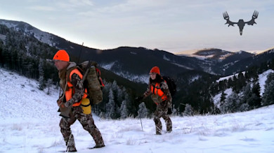 Two hunters in camouflage and blaze orange hiking up a snowy mountain slope, one carrying a rifle