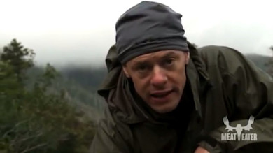 Steven Rinella leaning toward camera outdoors wearing bandana and rain jacket, MEAT EATER logo