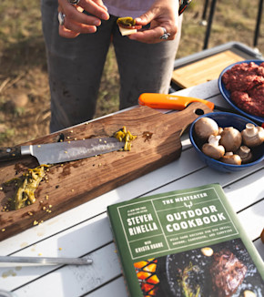 Book cover reads STEVEN RINELLA OUTDOOR COOKBOOK; hands prepping peppers on cutting board with mushrooms and raw meat