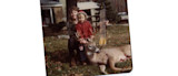 Man kneeling behind child in red coat holding buck antlers; bow and arrows beside deer on lawn