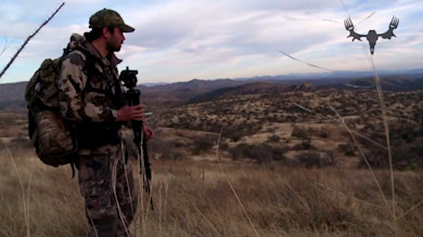 Hunter in camouflage holding tripod on hilltop overlooking scrubby hills, MeatEater logo