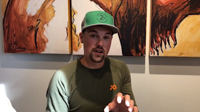 Man wearing green hat with text "2%" and First Lite shirt, gesturing before abstract paintings