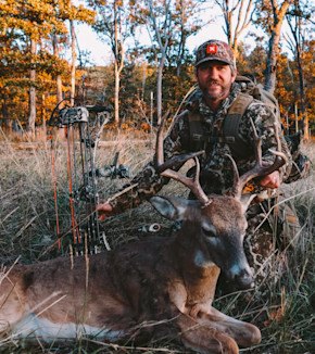 Clay Newcomb kneeling in camo holding a buck's antlers beside a compound bow
