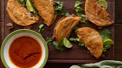 Crispy birria tacos on wooden board with cilantro, lime wedges and bowl of red dipping sauce