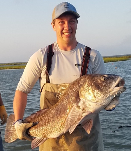 Man in cap and chest waders holding a very large fish in shallow marsh water