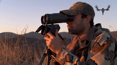 Hunter using binoculars on tripod at sunset, wearing camo cap and jacket