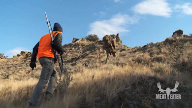 Two hunters climbing rocky hillside, one in orange vest carrying rifle; MEAT EATER logo