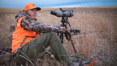 Hunter using spotting scope, seated in tall grass wearing orange vest and cap; rifle nearby