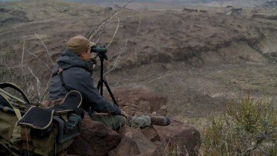 Hunter sitting on cliff, scanning rocky desert with spotting scope on tripod beside backpack.