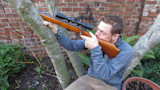 Man seated between tree trunks aiming a scoped rifle next to a brick wall