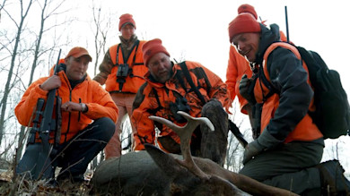 Five hunters in orange kneel around a harvested whitetail buck in bare winter woods