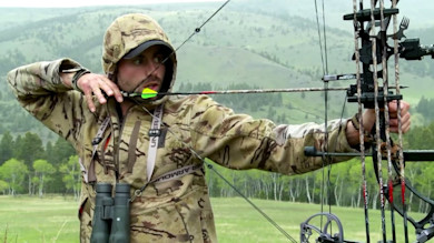 Hunter in camo hoodie drawing compound bow with green-fletched arrow, hilly meadow background