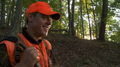Hunter smiling in orange cap and hunting vest on forested hillside at sunset