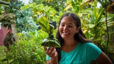Woman holding wrapped lau lau of taro leaves, smiling in a garden