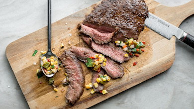 Chili-rubbed venison steak, sliced medium-rare, on wooden cutting board with corn salsa and carving knife