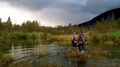 Two hunters seen from behind wading through shallow marsh carrying rifles toward tree line