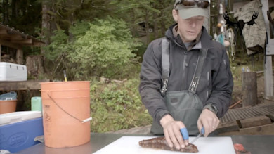 Man in cap and waders slicing a sea cucumber on a white cutting board outdoors