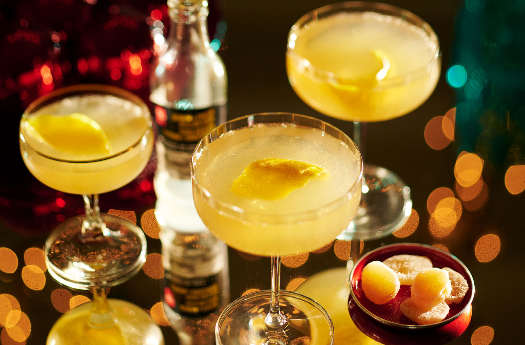 Glasses of cocktails on a table garnished with lemon slices next to a bottle of gin and bowl of candied ginger pieces