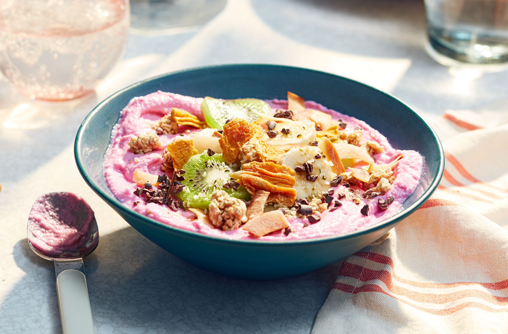 Une table avec un bol de smoothie au yogourt grec et aux fruits tropicaux garni de kiwi, de banane, de granola, de noix de coco et de graines de chanvre décortiquées.