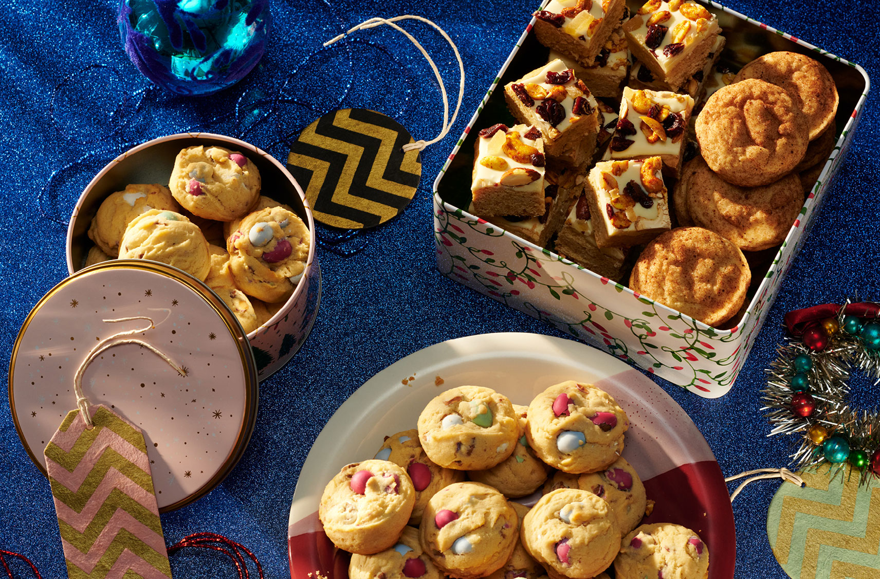 Une table remplie de boîtes de biscuits, dont des biscuits aux œufs en chocolat sur une assiette rouge et blanche à côté d'une boîte de snickerdoodles et de carrés garnis de cerises et de noix.