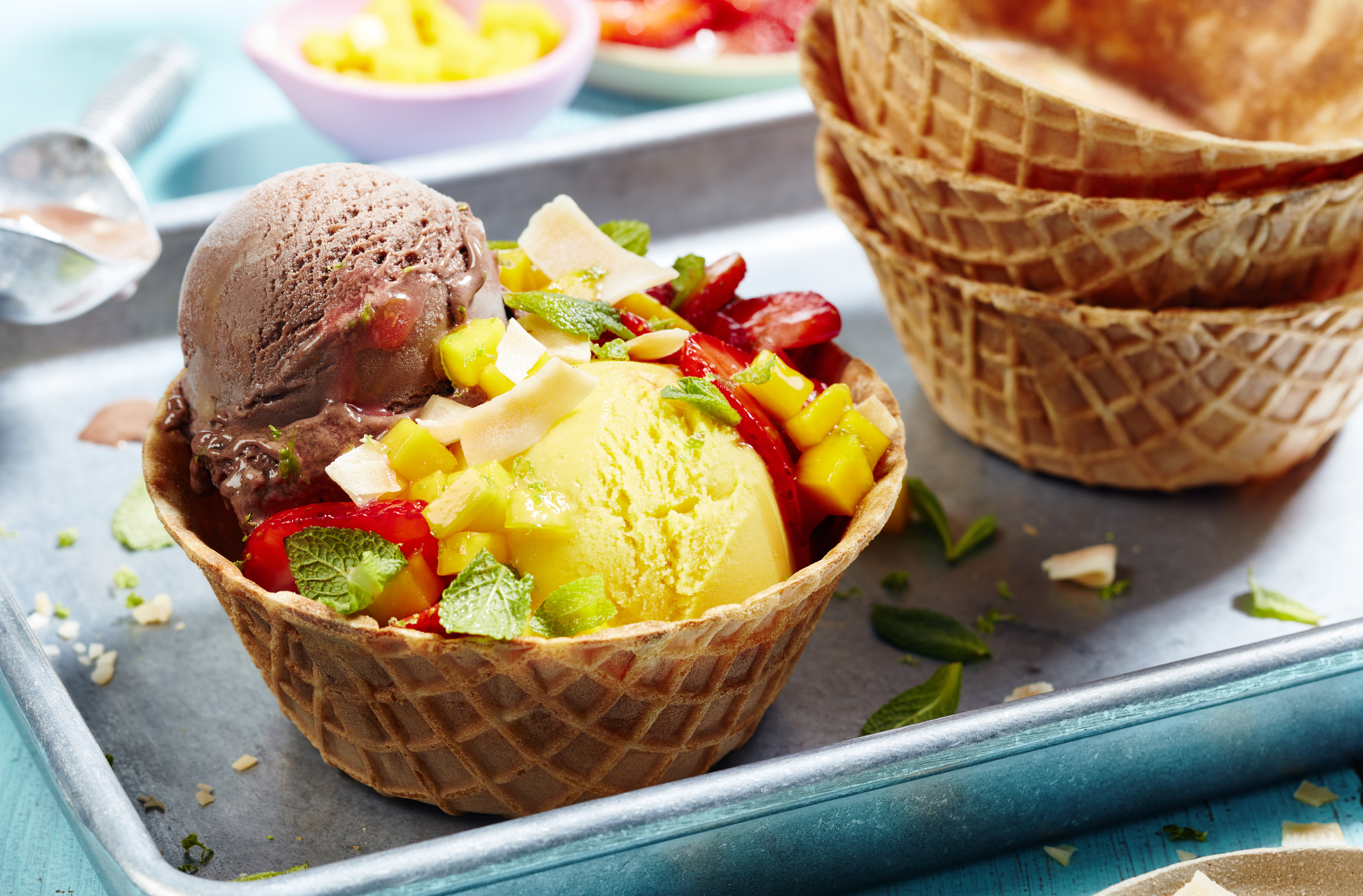 Dairy-Free chocolate and mango sundaes topped with chunks of mangoes, slices of strawberries and shavings of toasted coconuts. Mint leaves sprinkled on top. Served in waffle bowl